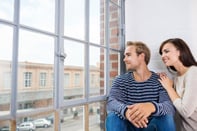Young couple watching life in the street below as they sit in front of a large window in their urban apartment